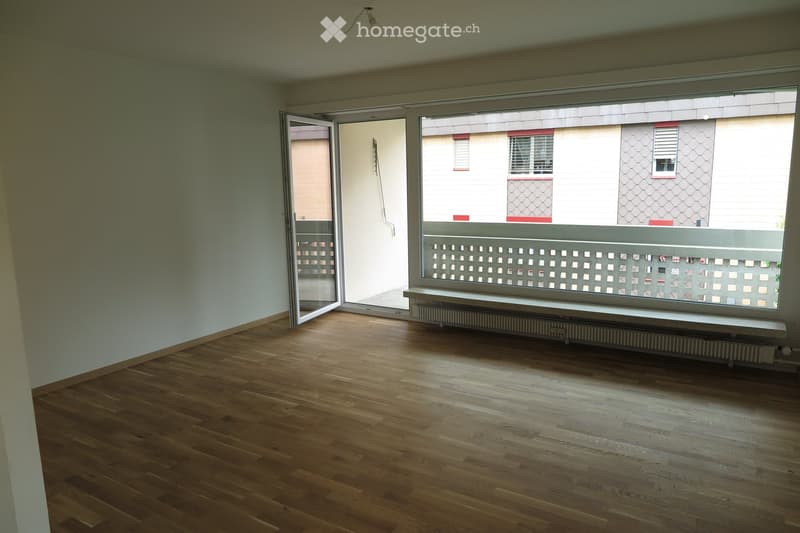 Empty room with wooden flooring, large window with a decorative white lattice, and a radiator below the window. The room has white walls and a sliding glass door leading to a balcony.