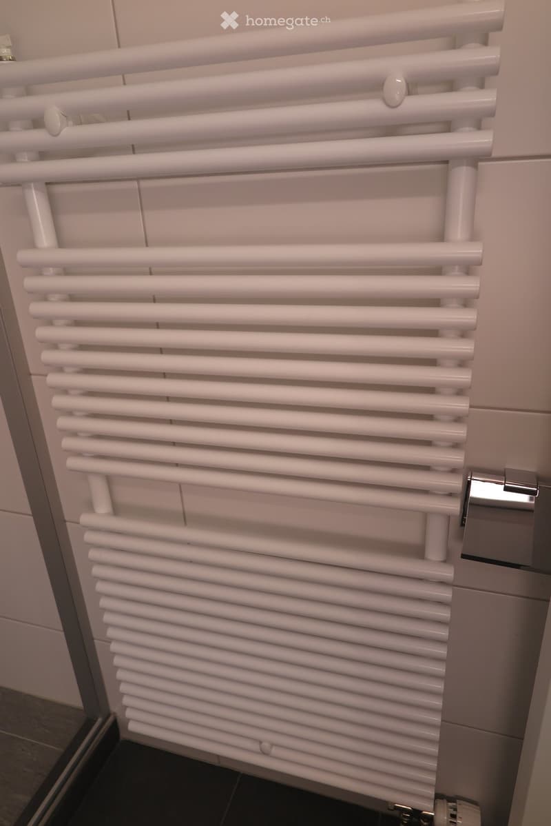 White radiator with multiple horizontal bars mounted on a tiled wall in a bathroom.