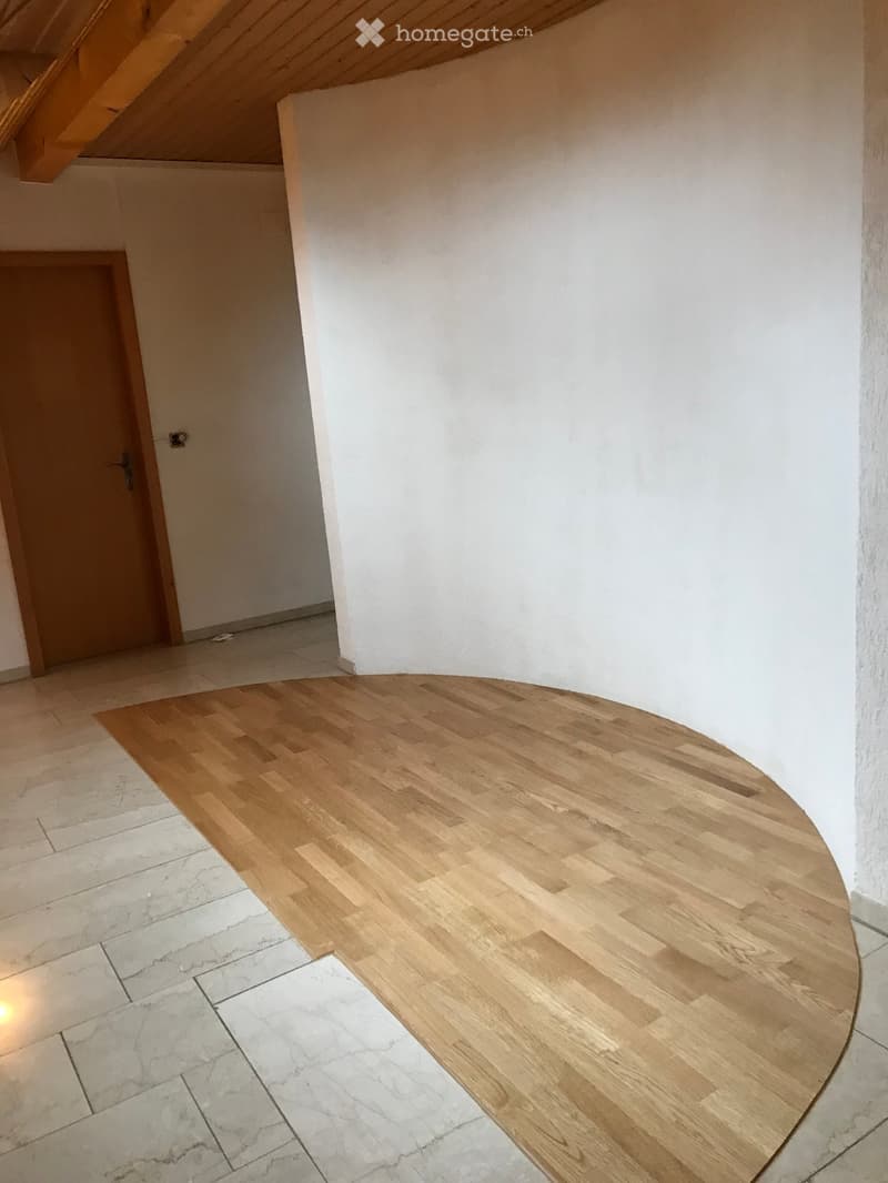 Empty room with wooden flooring and white walls, featuring a wooden door and a slanted wooden ceiling.
