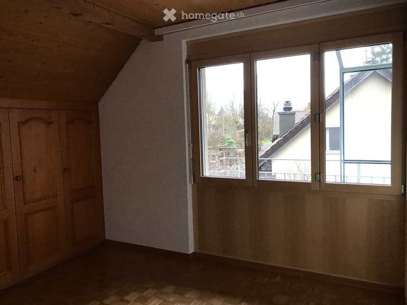 Empty attic room with wooden walls, slanted ceiling, large windows, and a wooden door.