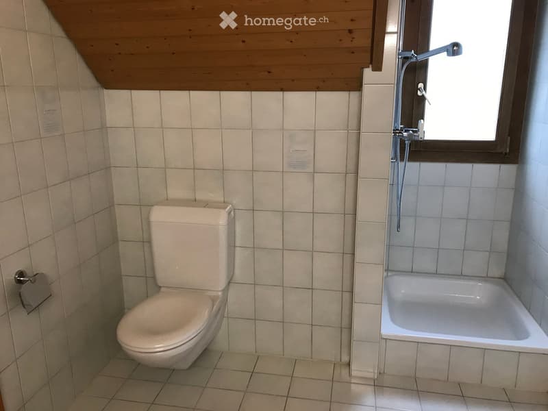 Small bathroom with a toilet, bathtub, and a window with a showerhead above it. The walls and floor are tiled in light beige.