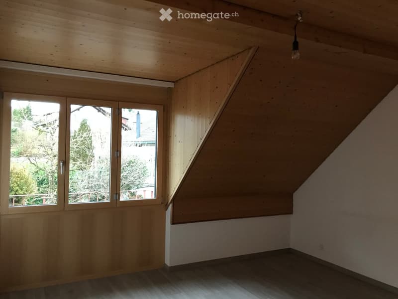 Empty attic room with wooden walls, slanted ceiling, large window, and wooden floor.