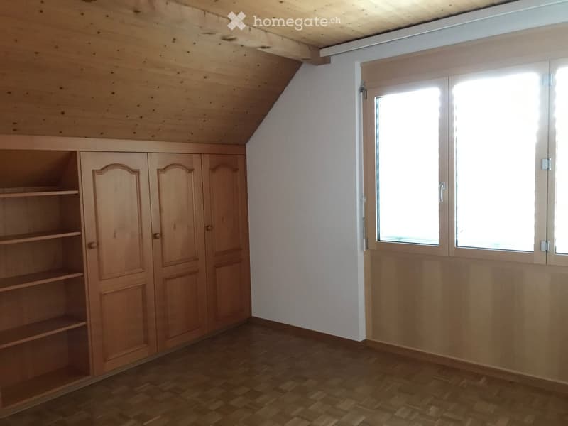 Empty attic room with wooden walls, slanted ceiling, large window, and built-in wooden cabinet with shelves.