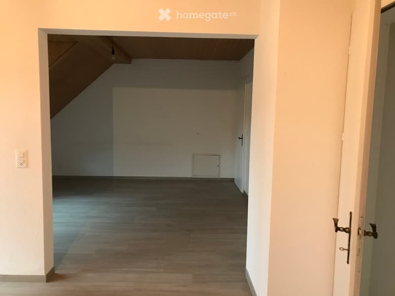Empty room with wooden flooring and white walls, featuring a doorway leading to another room with a slanted ceiling.