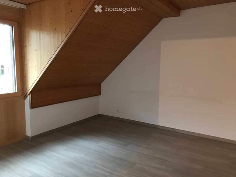 Empty attic room with wooden walls, sloped ceiling, and wooden flooring. Features a single window on one side and a small electrical outlet on the wall.
