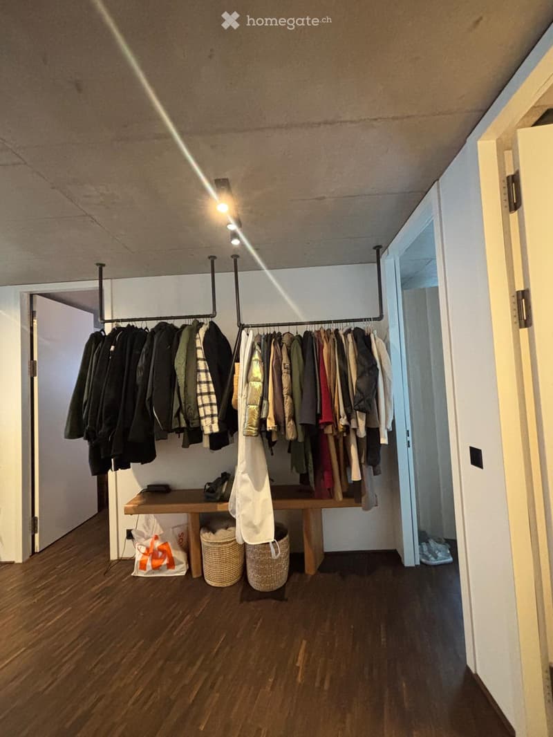 A well-organized walk-in closet with wooden flooring, a clothing rack holding various garments, a small wooden table with baskets and a handbag, and a few open doors leading to other spaces.