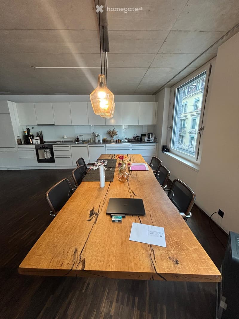 A modern, well-lit room with a large wooden table surrounded by black chairs, a laptop, papers, and a vase with flowers. The room features white cabinets, an oven, and a window with a city view.