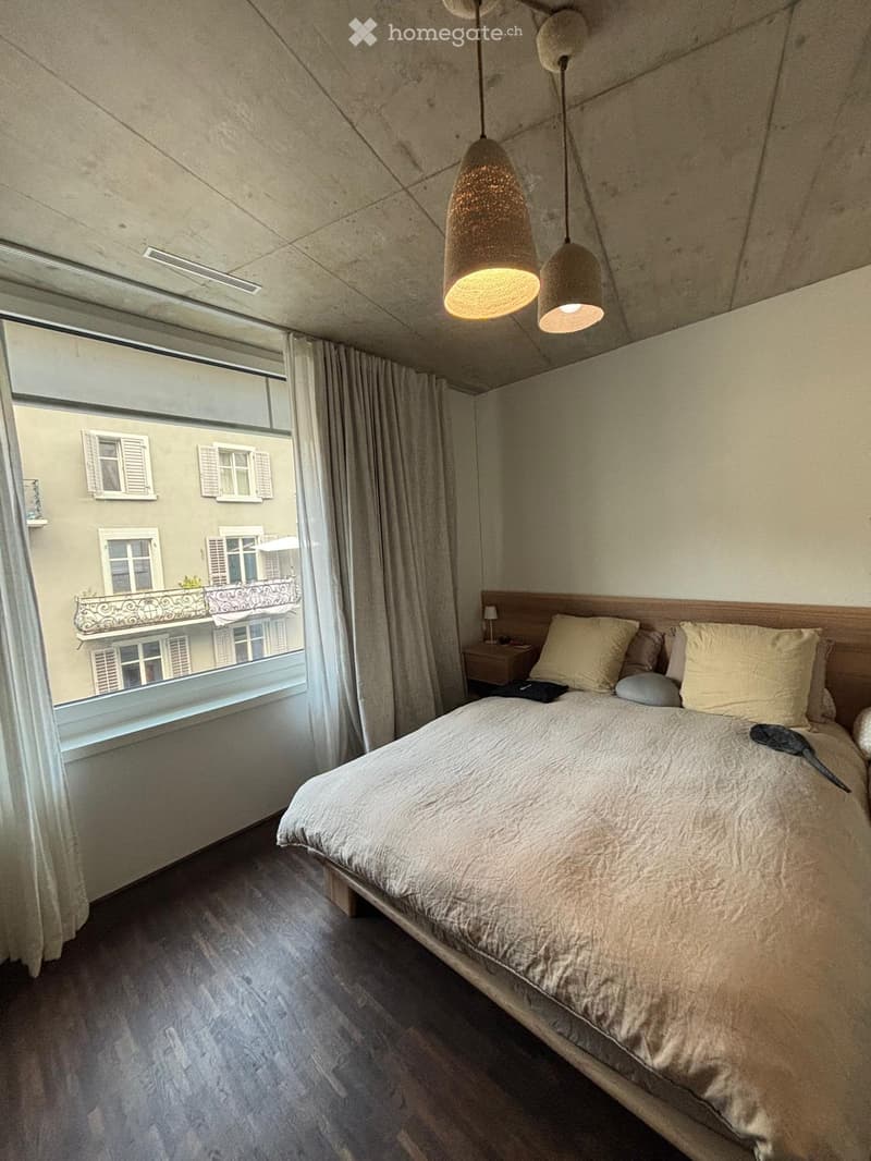 Modern bedroom with a large bed, nightstand, and large window with a view of a balcony on an adjacent building.