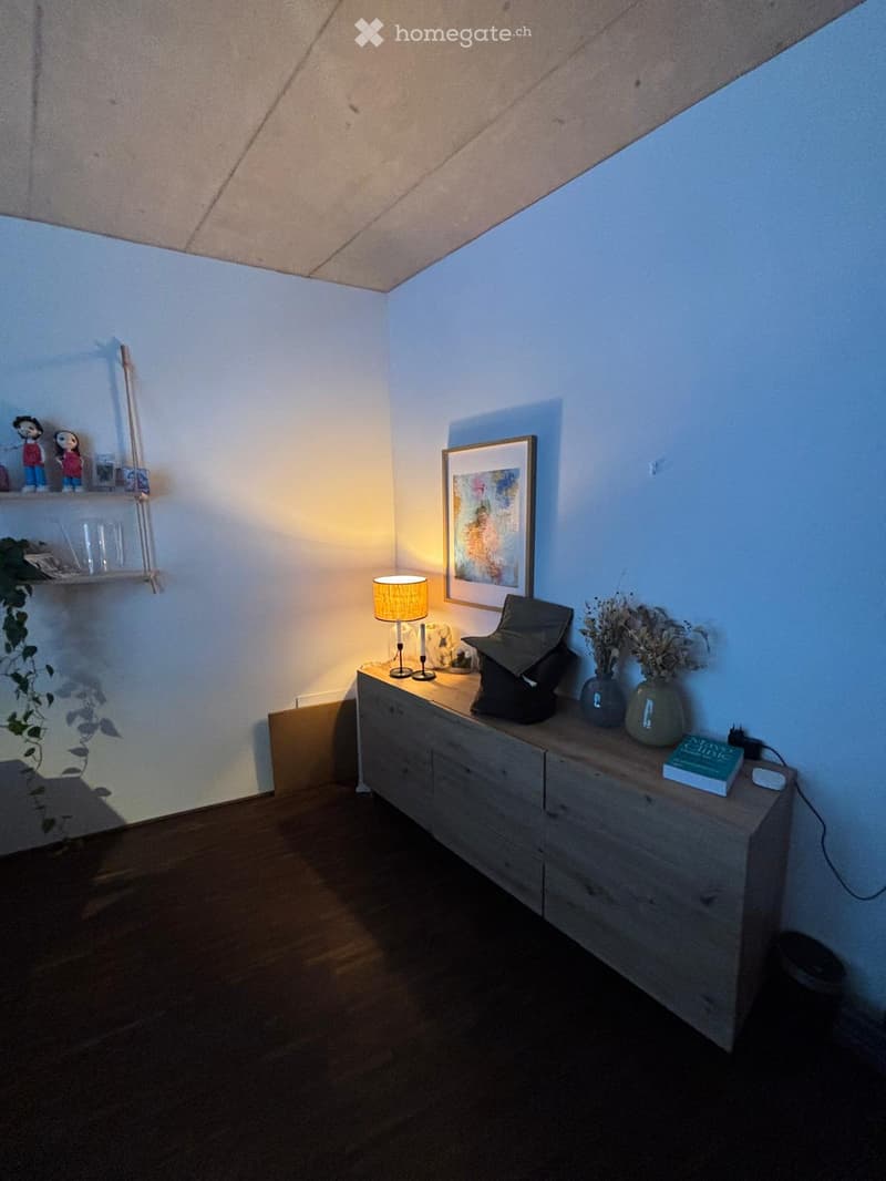 A small, well-lit room with a wooden table, a lamp, vases, a picture frame, a shelf with decorative items, and a plant. The room has a slanted ceiling and wooden flooring.