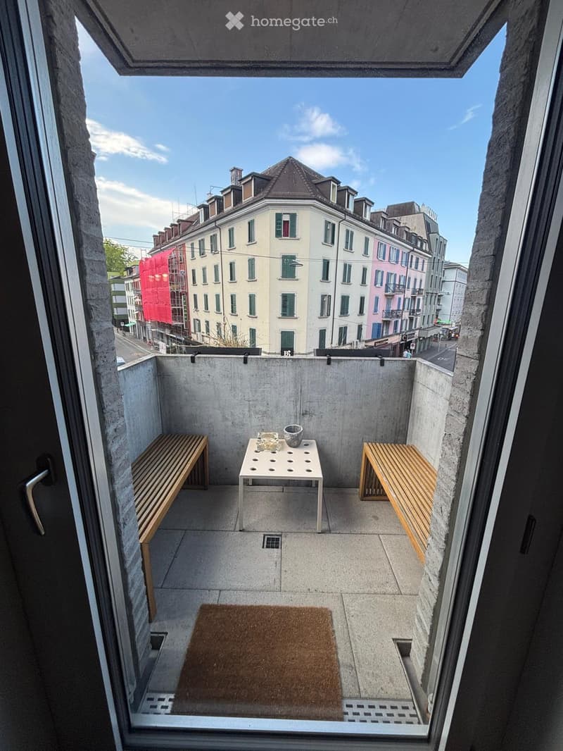 Small concrete balcony with a view of the city, featuring a white table with a vase, two wooden benches, and a brown doormat.