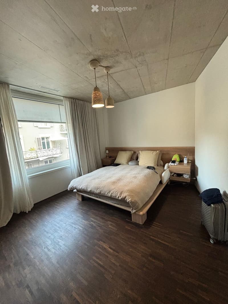 Modern bedroom with wooden floor, large window with curtains, bed with pillows and duvet, nightstand with lamp, and hanging pendant lights.