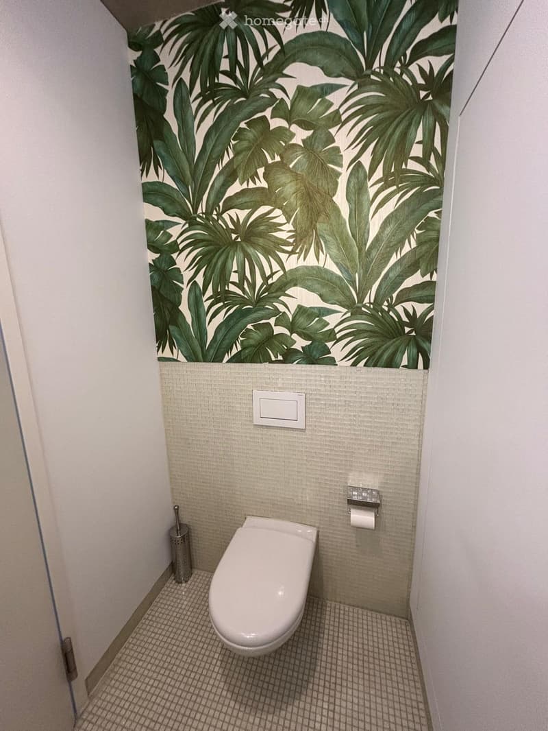 Modern bathroom with a white toilet, beige tiled walls, and a decorative leafy green wallpaper accent above the tiles.