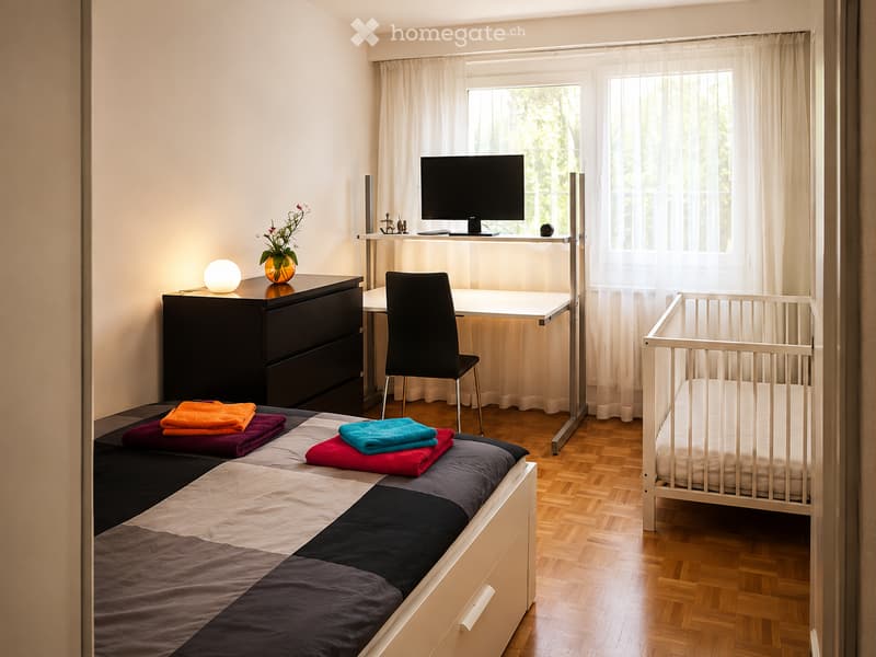 A well-lit bedroom with a single bed, desk, chair, and a small cradle. The room features a modern design with a wooden floor, large windows with sheer curtains, and decorative items including a flower vase and a lamp.