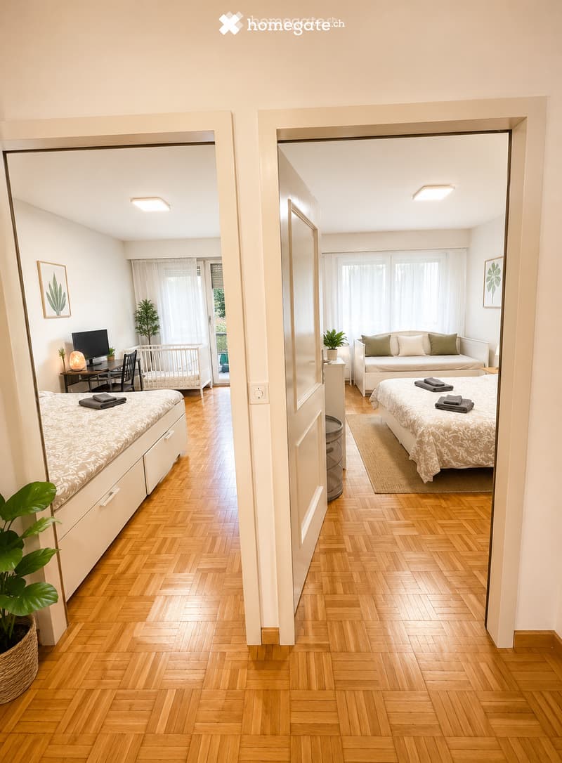 A well-lit, modern furnished room with wooden flooring, a bed, a sofa, a desk with a screen, and a small plant. The room features large windows with white curtains and a view of greenery outside.