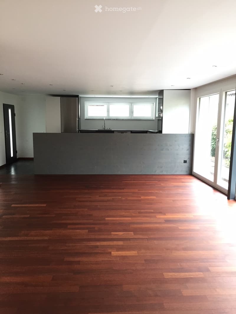 Empty room with wooden flooring, large windows, and a kitchen area visible in the background.
