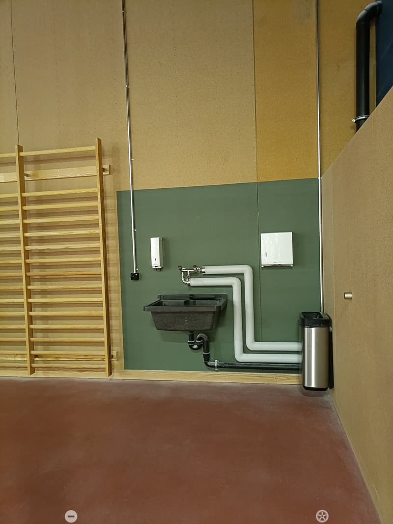 Bathroom with a black sink, faucet, soap dispenser, paper towel dispenser, and a trash can. Wooden shelf on the left side.