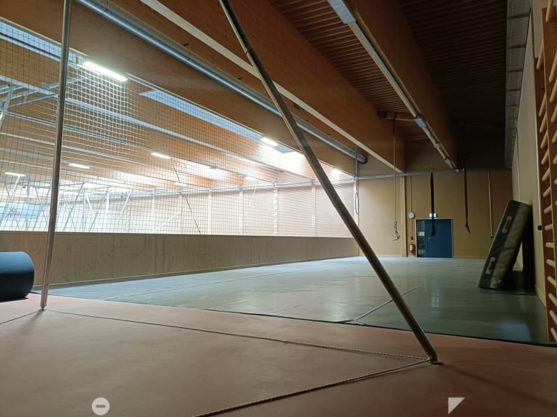 Large, open gymnasium with wooden beams, large windows, and a padded floor. No furniture or equipment is visible.