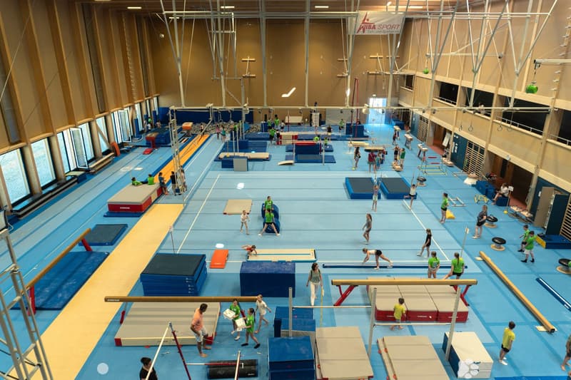 A spacious indoor gymnastics training facility with multiple padded exercise areas, balance beams, uneven bars, and a vaulting table. Young gymnasts and coaches are practicing various routines.