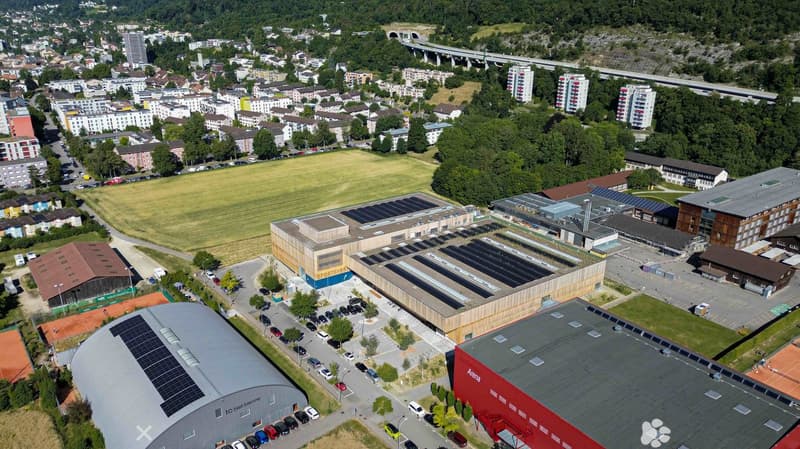 Aerial view of a modern sports complex with multiple buildings, solar panels on roofs, parking lots, and surrounding urban area with residential buildings and green spaces.
