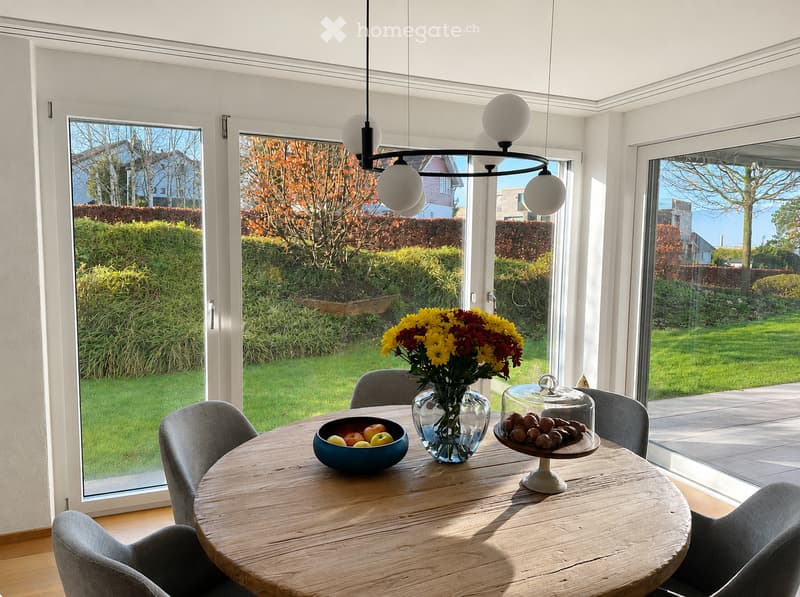 Bright, modern kitchen with large windows overlooking a garden, featuring a round wooden dining table with chairs, a vase of flowers, a bowl of fruit, and a cake on display.
