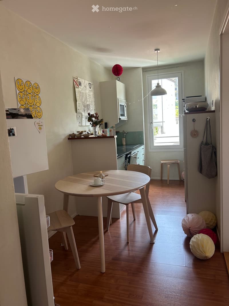 A modern, compact kitchen and dining area with wooden flooring, a round wooden dining table with four chairs, a small kitchen counter with a microwave, and a refrigerator. Decorative balloons and a vase with flowers are visible.