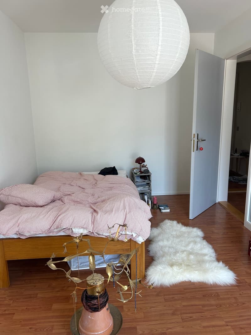 Modern bedroom with wooden flooring, a bed with pink striped bedding, a nightstand with a table lamp, a shaggy white rug, and a decorative sculpture on the floor.