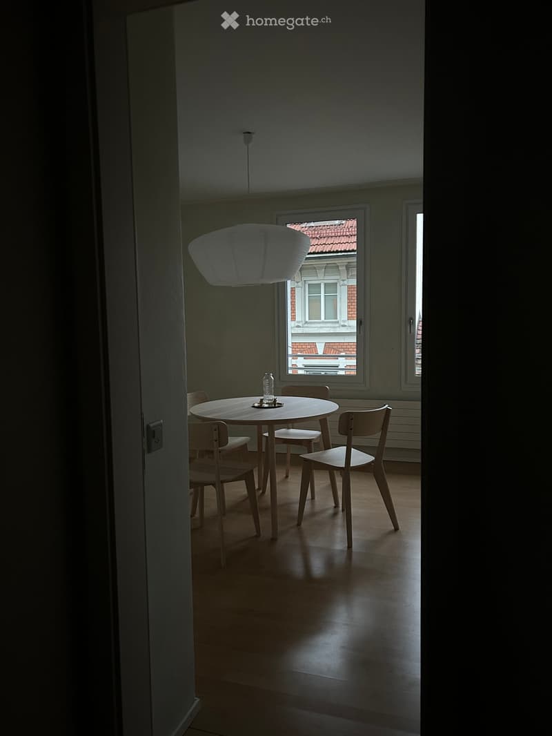 Dining area with a round wooden table, four matching chairs, and a modern pendant light hanging above. The room has wooden flooring and large windows showing a view of a neighboring building.