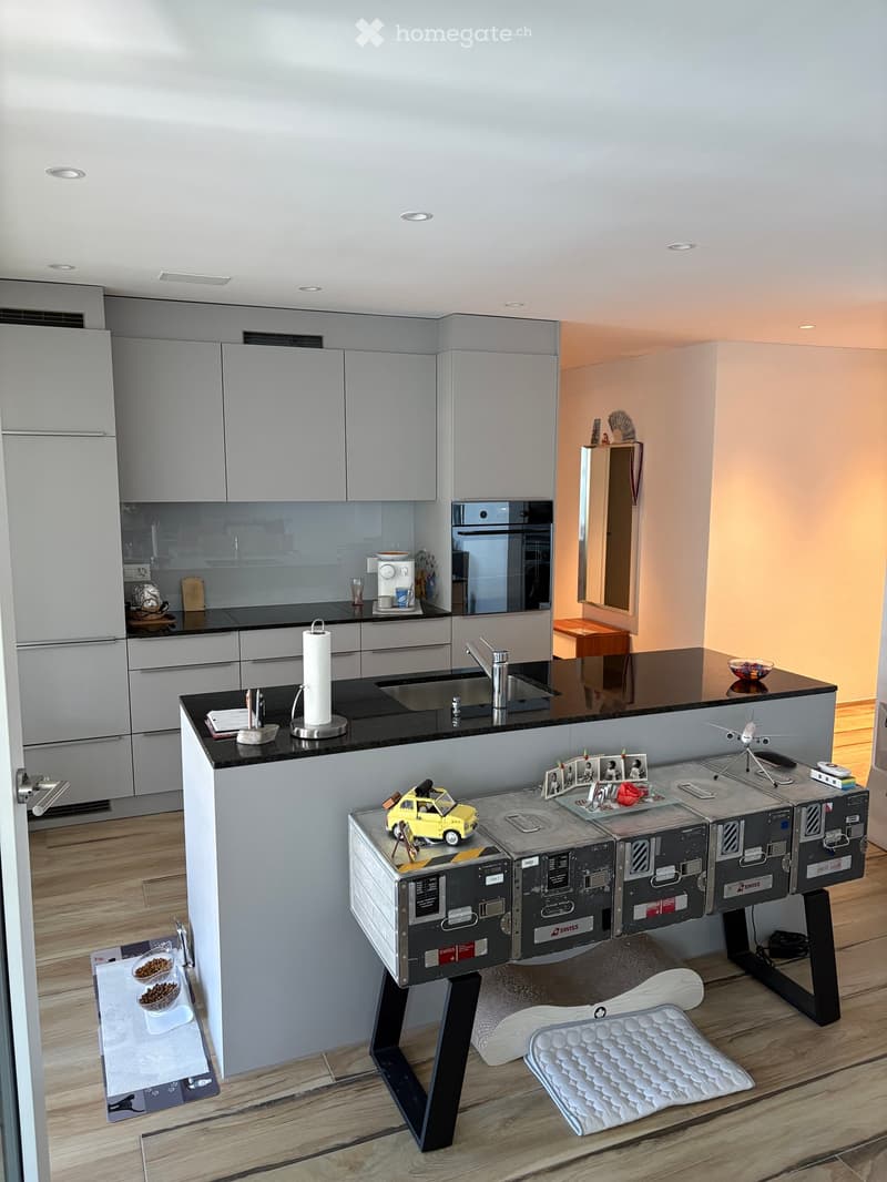 Modern kitchen with grey cabinets, black countertops, a sink, coffee maker, and a small seating area with a stool and chair.