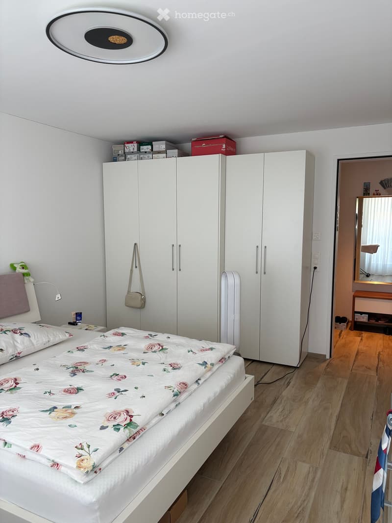 Modern bedroom with a white bed, floral duvet, white wardrobe, mirror, and a small stool in the adjacent room.