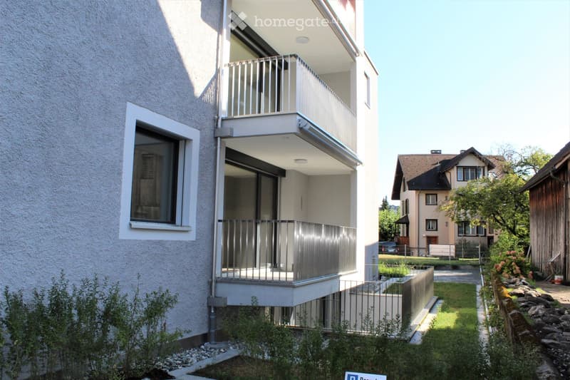 Modern two-story building with a balcony, surrounded by greenery and neighboring houses.