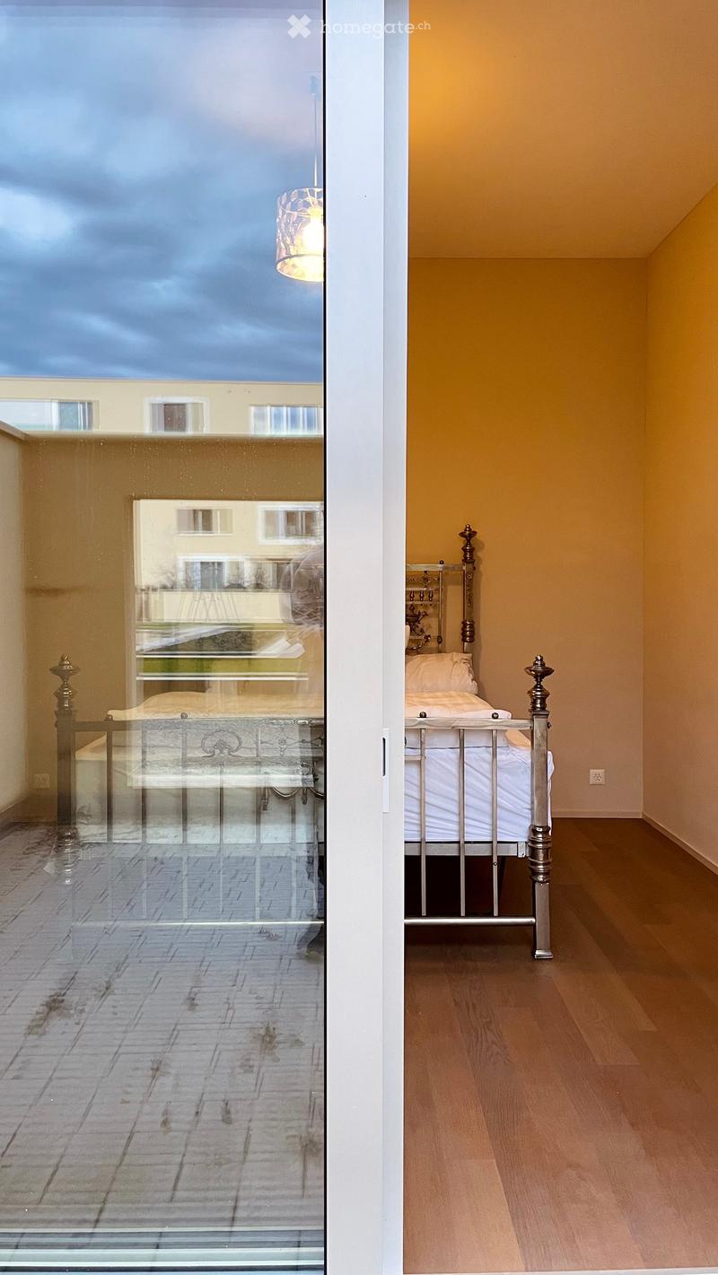Cozy bedroom with a wooden floor, a metal-framed bed, and a lamp. A large window offers a view of a brick-paved outdoor area.