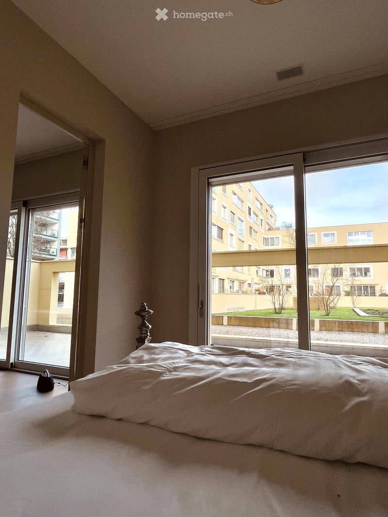 Bright bedroom with large windows and sliding doors leading to a balcony, featuring a neatly made bed and a decorative lamp.