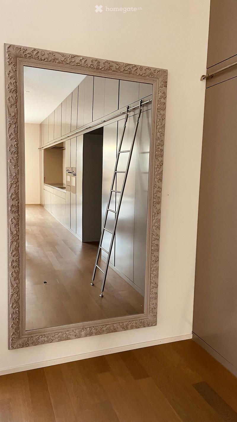 Empty room with light wooden flooring, white walls, and a large ornate mirror on one wall. A ladder is positioned in front of the mirror, and a kitchen area is visible in the background.