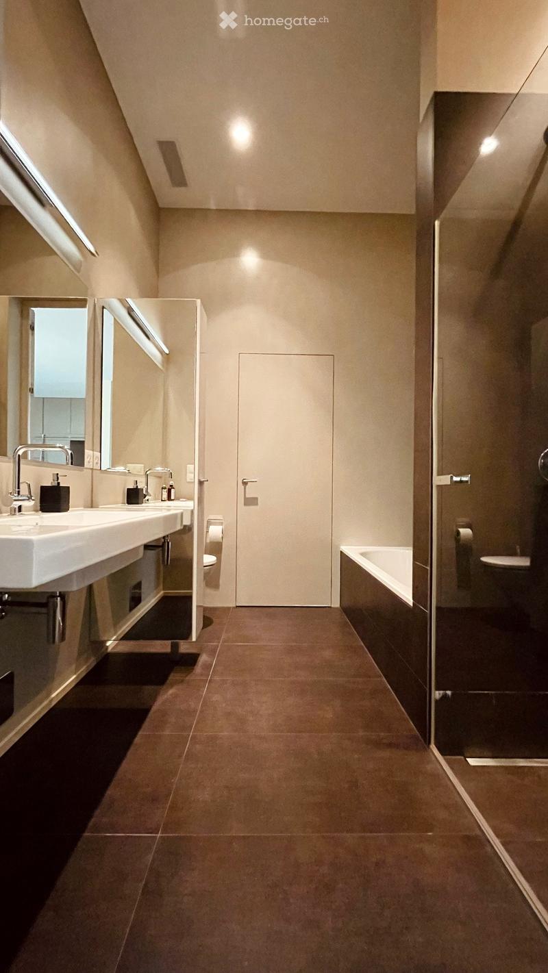 Modern bathroom with double sink, bathtub, toilet, and large mirror. Brown tiled floor and beige walls.