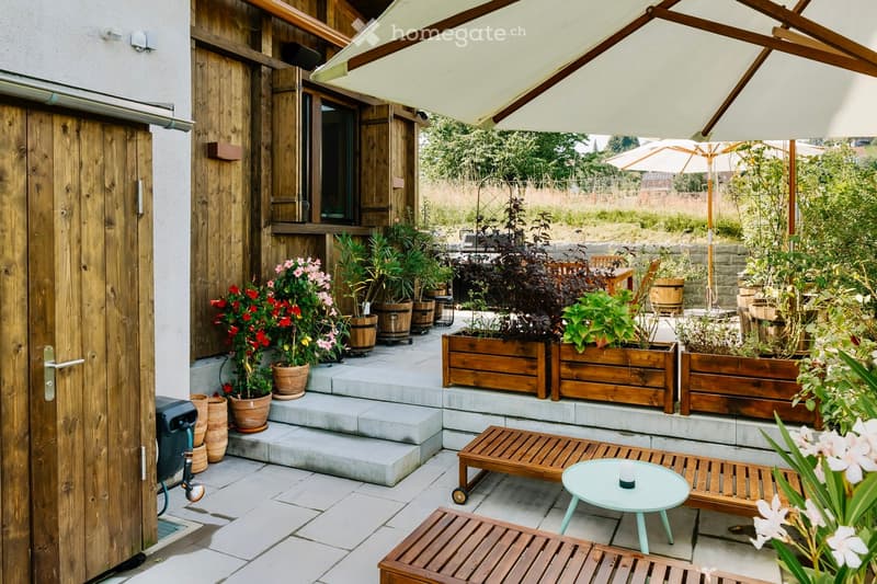 Outdoor patio area with wooden bench, green plants, flowers, umbrellas, table, and wooden doors.
