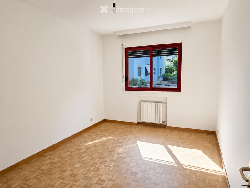 Empty room with wooden herringbone flooring, white walls, a large window with red frame, radiator, and a hanging light fixture.
