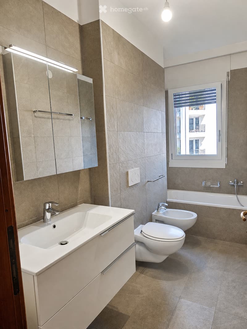 Modern bathroom with beige tiles, a double sink, toilet, bidet, bathtub, and a large mirror with built-in lighting. The room features a window with blinds and a sleek, contemporary design.