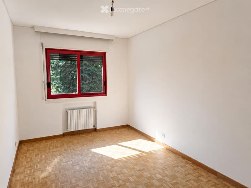 Empty room with white walls, wooden parquet flooring, a single window with red frame, radiator, and a hanging light fixture.