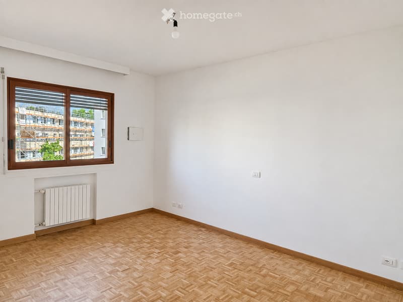 Empty room with wooden herringbone flooring, white walls, and a large window with wooden frame. Features a radiator below the window and a single ceiling light fixture.