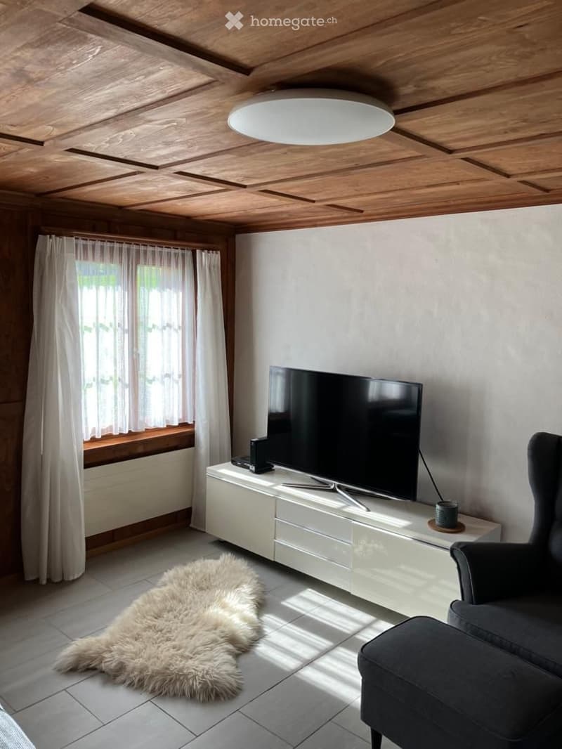 Modern living room with a large flat-screen TV on a white TV stand, a plush white rug, and a comfortable dark grey armchair. The room features wooden ceiling beams and a round ceiling light.