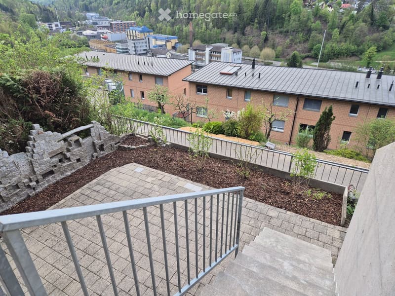 Balcony with tiled floor, metal railing, small landscaped area with plants and a stone feature, overlooking a residential area with buildings and greenery.