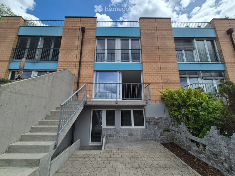Modern multi-story building with brick facade, blue accents, and balconies accessible via a raised ground floor staircase.