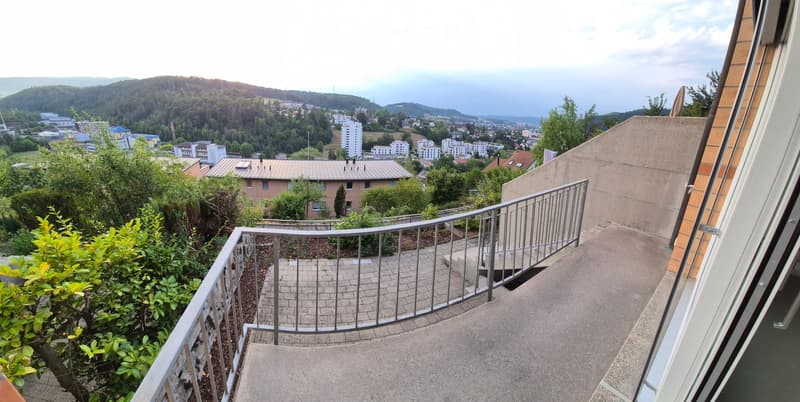 Balcony with metal railing, overlooking a cityscape with buildings and greenery.