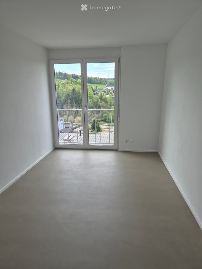 Empty room with light grey walls and carpeted floor, featuring a large sliding glass door leading to a balcony with outdoor furniture visible.
