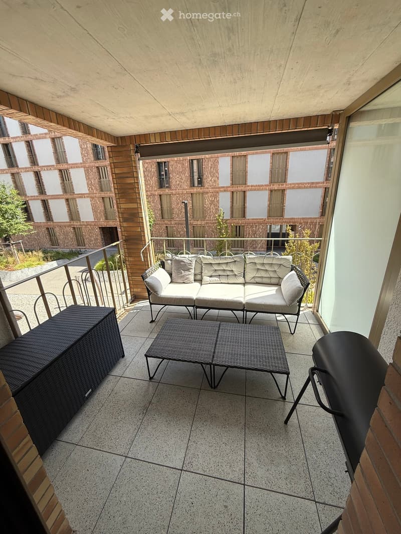 Balcony with a modern seating area, including a sofa, a small table, and a storage box, overlooking a building with brick facade and windows.