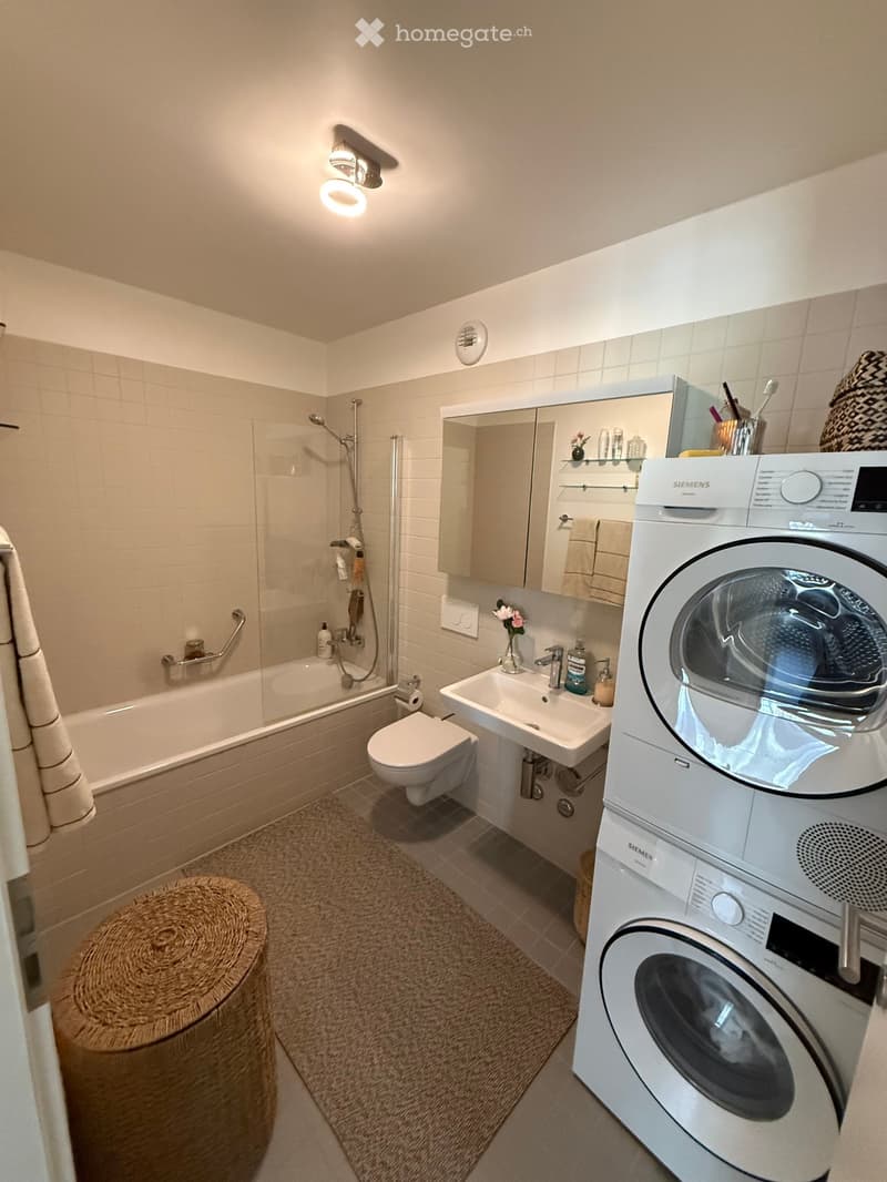 Modern bathroom with a bathtub, toilet, sink, mirror, and a stacked washing machine. Features include a wicker stool, a showerhead, and a towel hanging on the side.