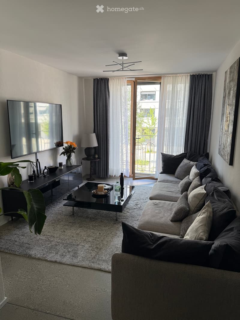 Modern living room with a large sectional sofa, coffee table, side table with lamp, television mounted on the wall, and a view to a balcony through large windows.