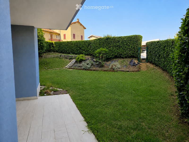 Well-maintained garden with a lawn, small plants, and a pathway leading to a house entrance. The area is bordered by hedges and includes a small staircase leading to a raised section.