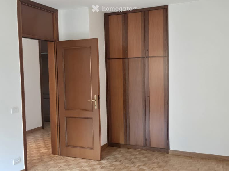 Empty room with wooden flooring, white walls, and a large wooden wardrobe. An open door leads to another room.