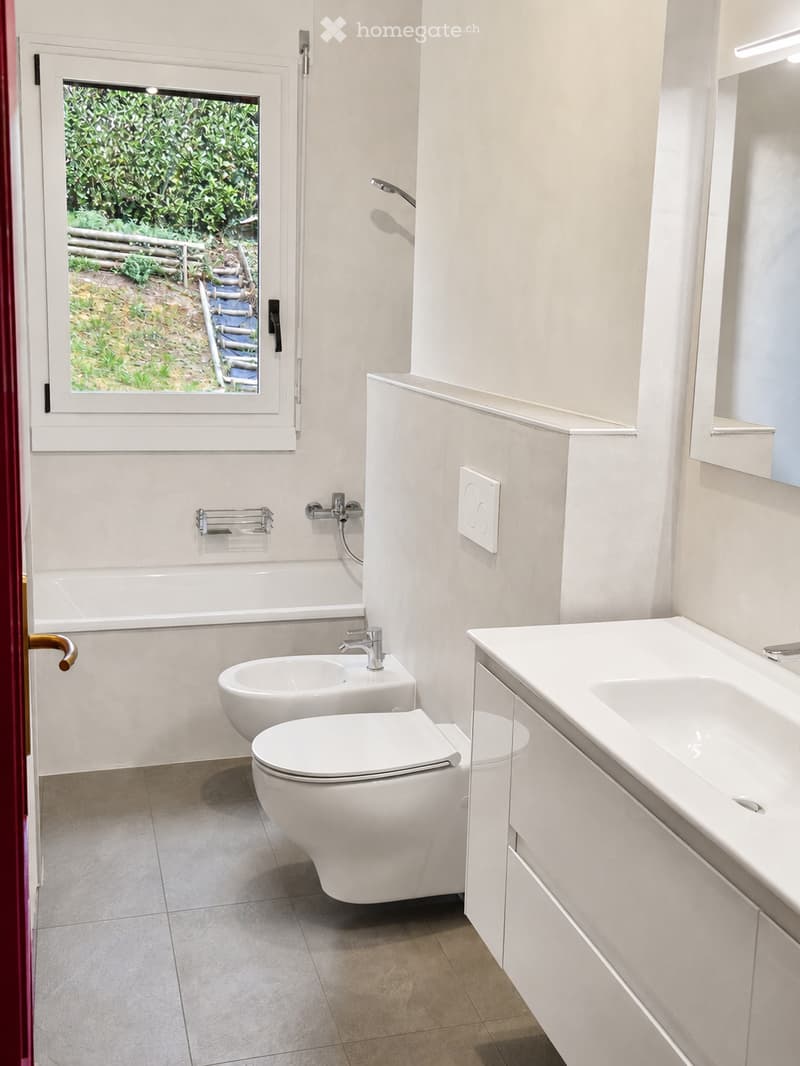 Modern bathroom with white fixtures, including a bathtub, toilet, sink, and a large window overlooking a garden.